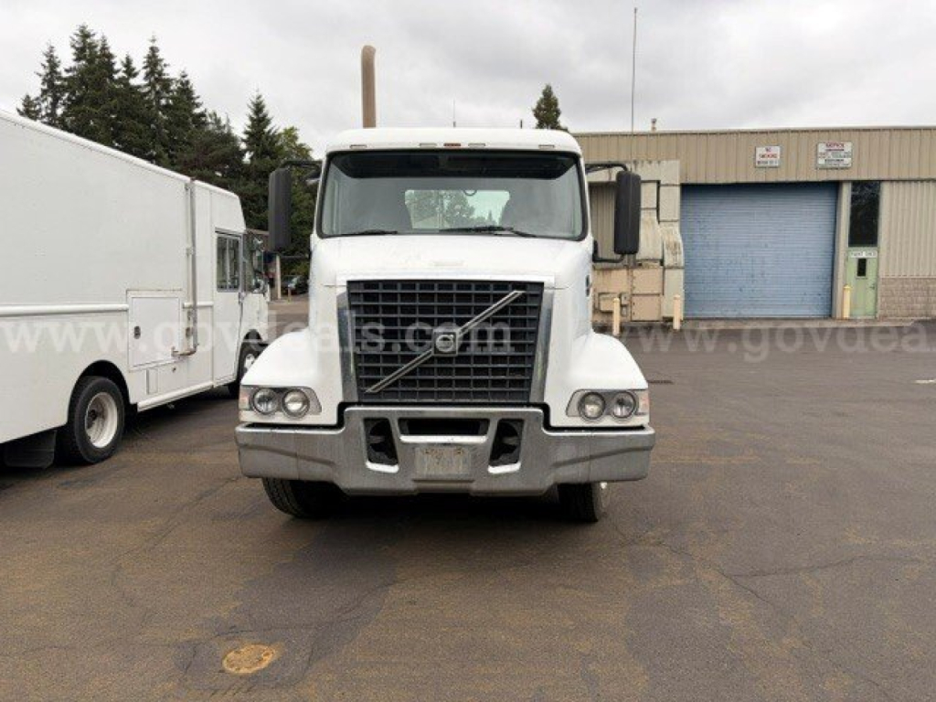 Front view of a white 2011 Volvo VHD truck with large grille, bumper, and headlights; two side mirrors; parked in an industrial lot.