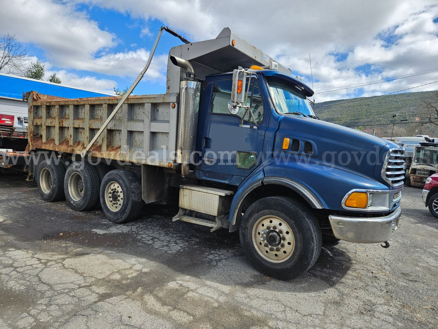 Dump truck showing blue Cab with gray dump bed, three rear axles, side steps, and front chrome bumper. Visible text on image includes www.govdeals.com watermark.