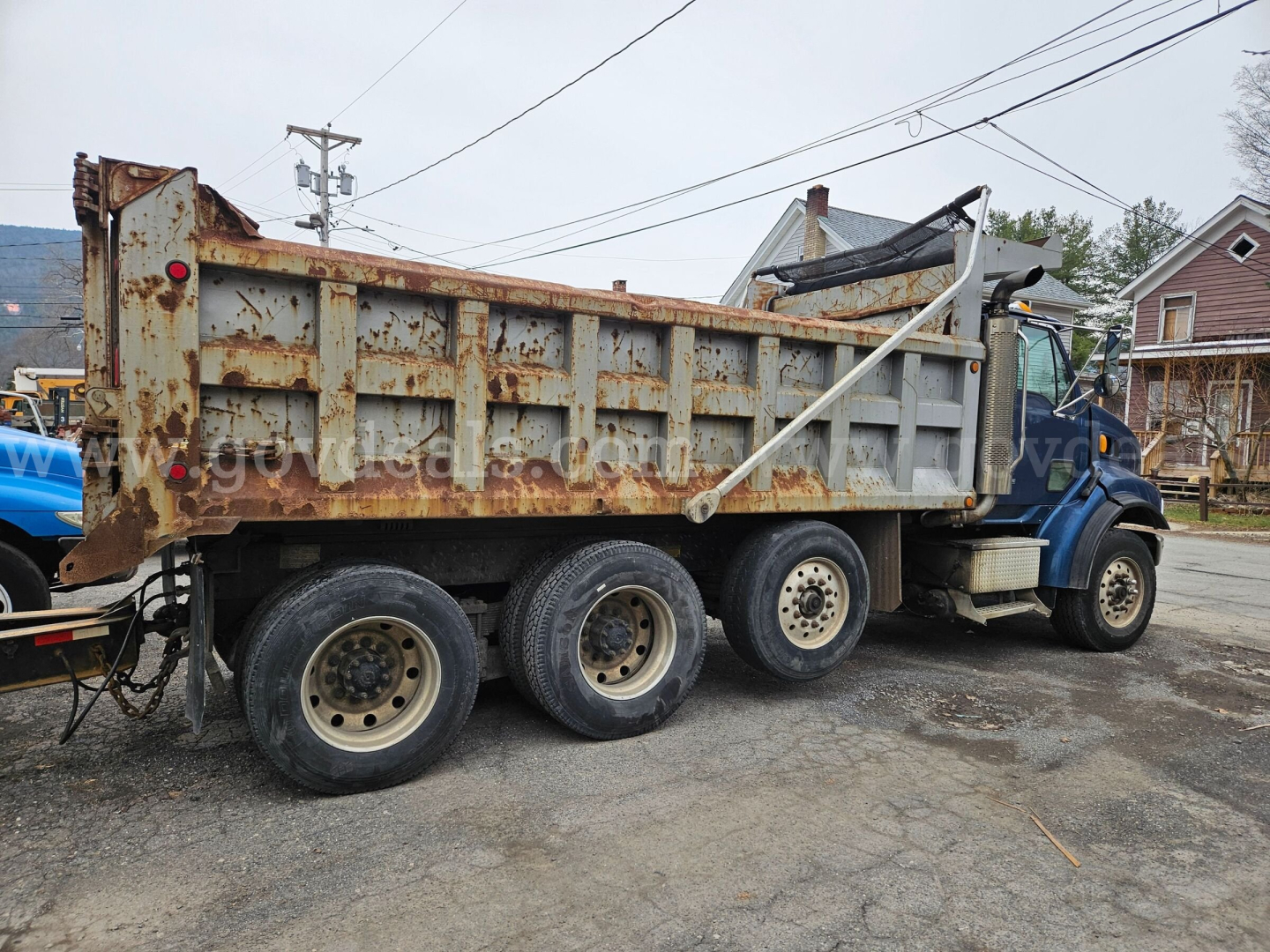 Dump truck side view showing rusted steel dump bed with three rear axles and blue cab, visible weathered hopper and support bars.