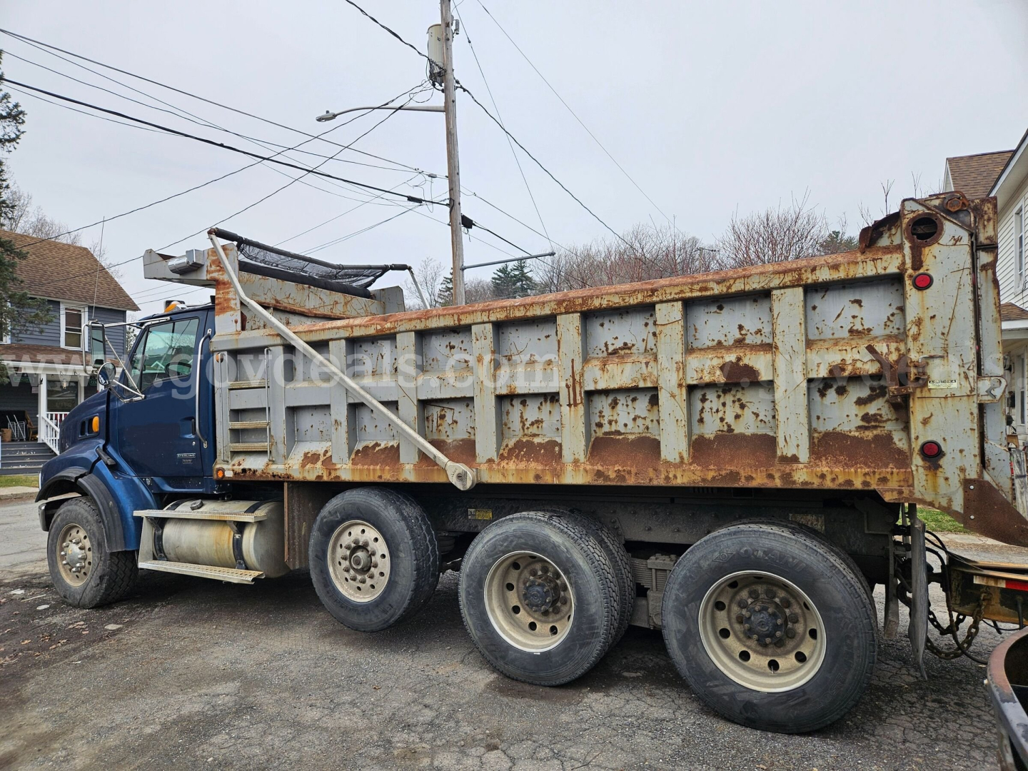 Dump truck side view showing a blue cab and a rusted dump body with six wheels, carrying a worn metal bed and hydraulic mechanism under the cab.