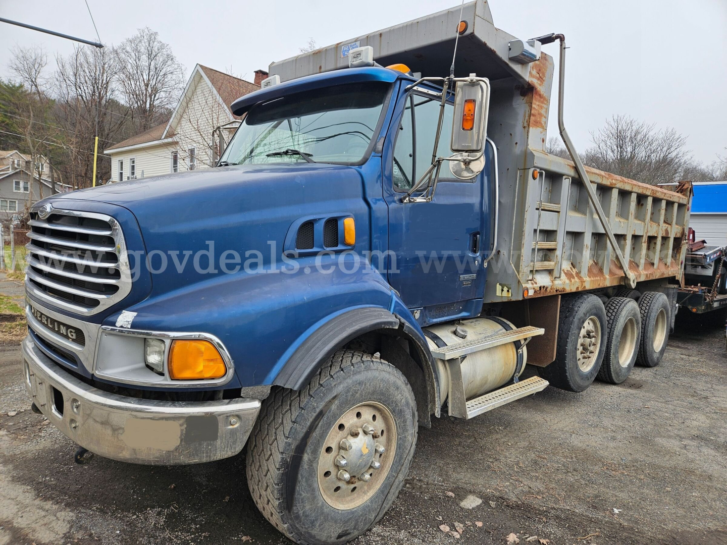 Dump truck showing blue Sterling LT9500 cab and gray dump bed with rust. Dual rear axles, front grille, headlights, and large side mirror visible.