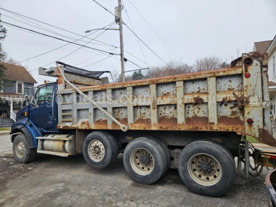 Dump truck side view showing a blue cab and a rusted dump body with six wheels, carrying a worn metal bed and hydraulic mechanism under the cab.