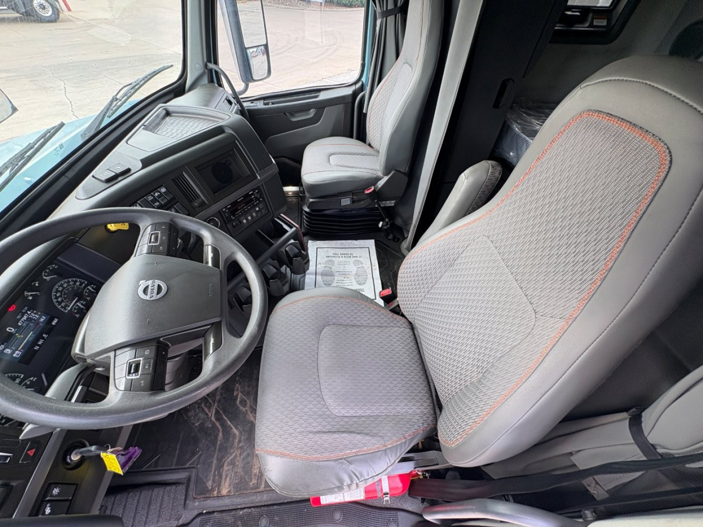 Truck cab interior visible: steering wheel with Volvo logo, dashboard, driver seat with gray fabric and orange piping, passenger seat, floor mat, paperwork on the floor. No legible labels or stickers readable.