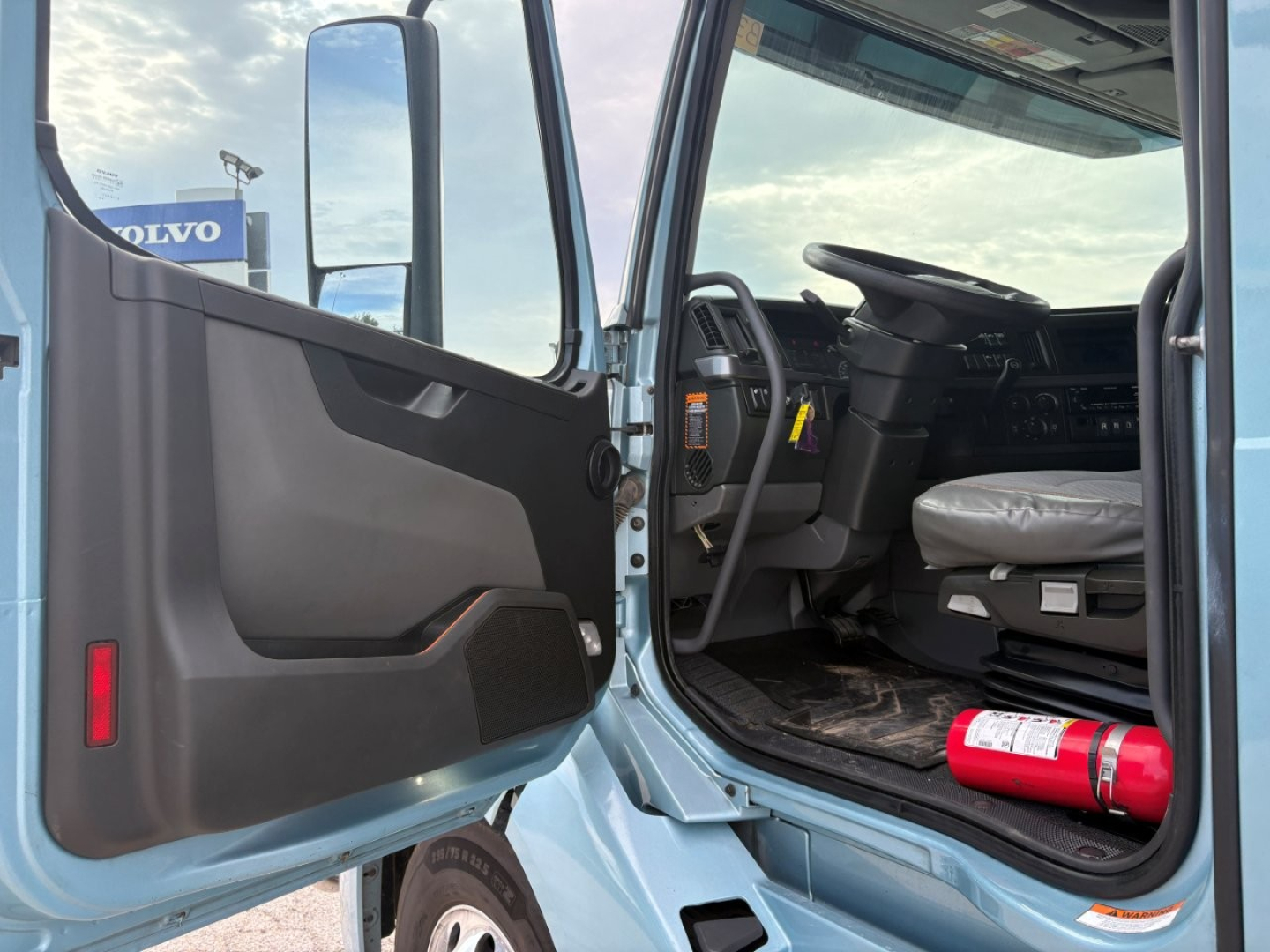 Volvo VNL64T760 truck cab interior with open door showing steering wheel and driver's seat, dashboard, and a red fire extinguisher on the floor؛ Volvo dealership signage visible outside window.