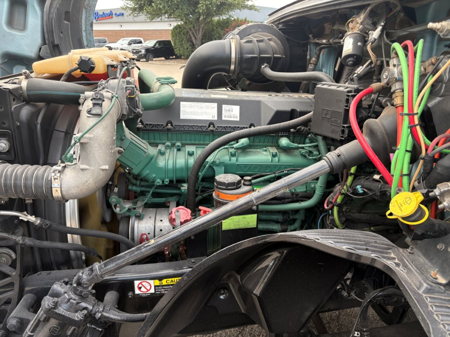Volvo engine bay showing green D13 engine with various hoses, wiring, and a yellow oil cap; red, green, and black cables near the firewall; side view of the turbo intake and components.