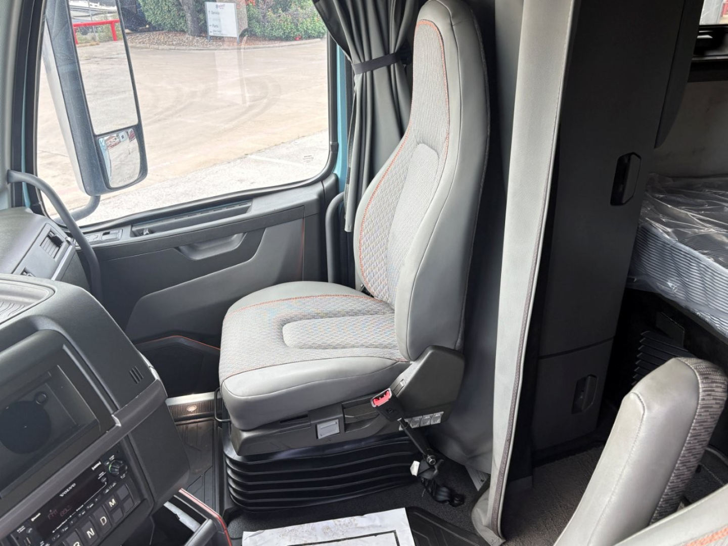 Interior of a Volvo VNL64T760 truck cab showing driver seat and passenger seat with gray fabric, dashboard, and side window; part of sleeper area visible.