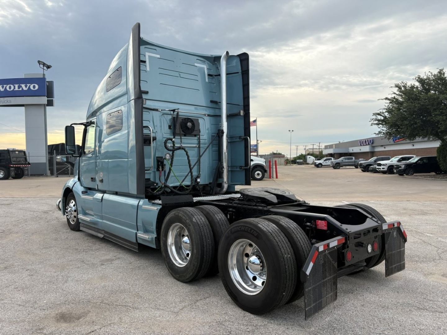 Volvo VNL64T760 truck in a dealership lot, rear view showing sleeper cab, dual rear axles, and fifth wheel. Visible text includes Volvo sign in background.