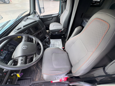 Truck cab interior visible: steering wheel with Volvo logo, dashboard, driver seat with gray fabric and orange piping, passenger seat, floor mat, paperwork on the floor. No legible labels or stickers readable.