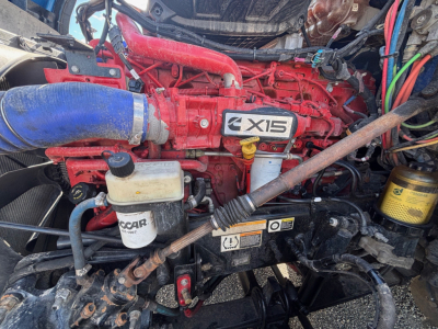 Peterbilt 579 engine bay showing Cummins X15 engine with red block, blue intercooler hose, reservoir bottles, and various hoses and wiring.