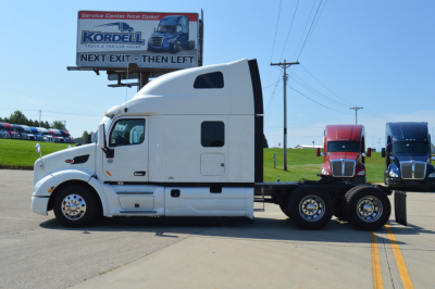 Peterbilt 579 semi-truck tractor cab side view, white paint, sleeper cab, with attached 2-axle trailer chassis visible; dealership sign in background.