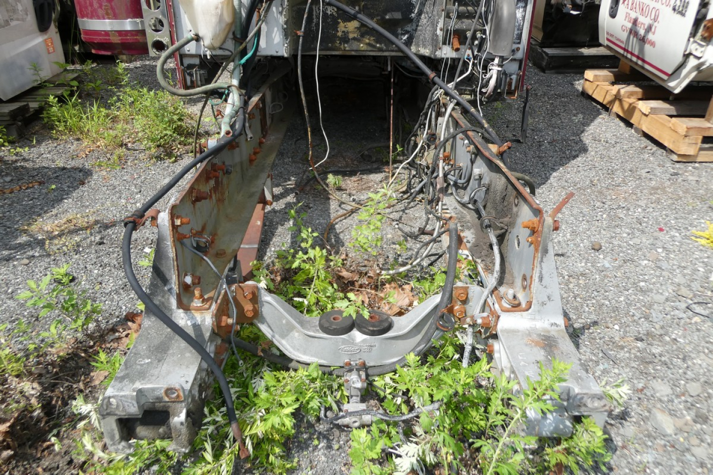 Vehicle chassis view showing front cab mount/frame with rust-free metal edges and overgrown weeds; engine bay area missing components and wiring harnesses visible.