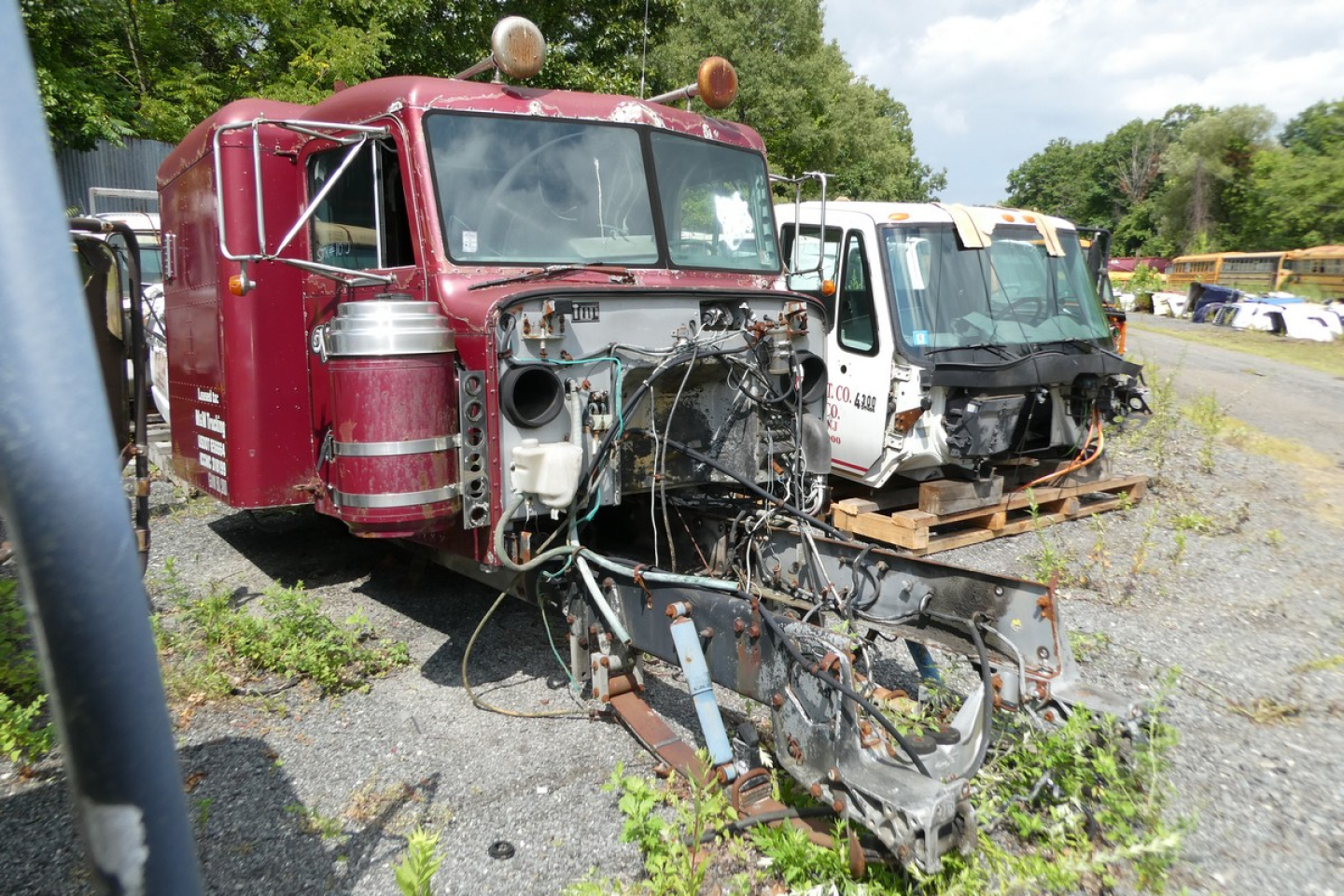 Peterbilt cab chassis in a salvage lot with exposed engine bay and wiring harnesses, front view, red paint, other cab units in background.
