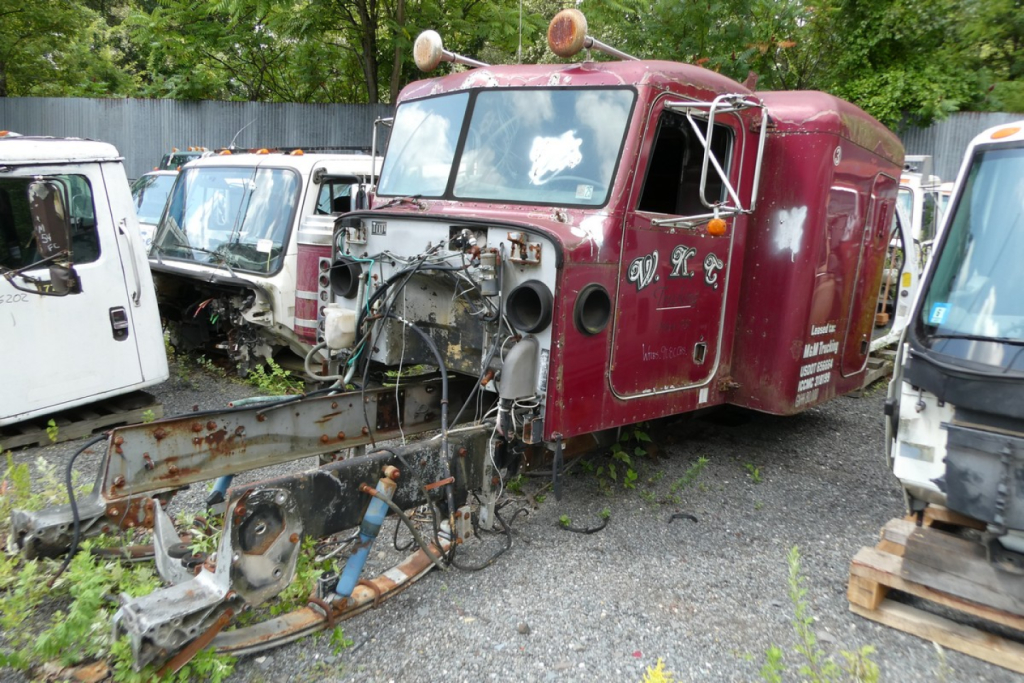 Peterbilt 379 cab in salvage yard showing exposed chassis and damaged front; maroon body with white lettering on cab doors.