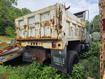Dump truck bed of an International 1854 with rusty steel dump body, rear view showing tailgate and mud flaps, overgrown vegetation around wheels.