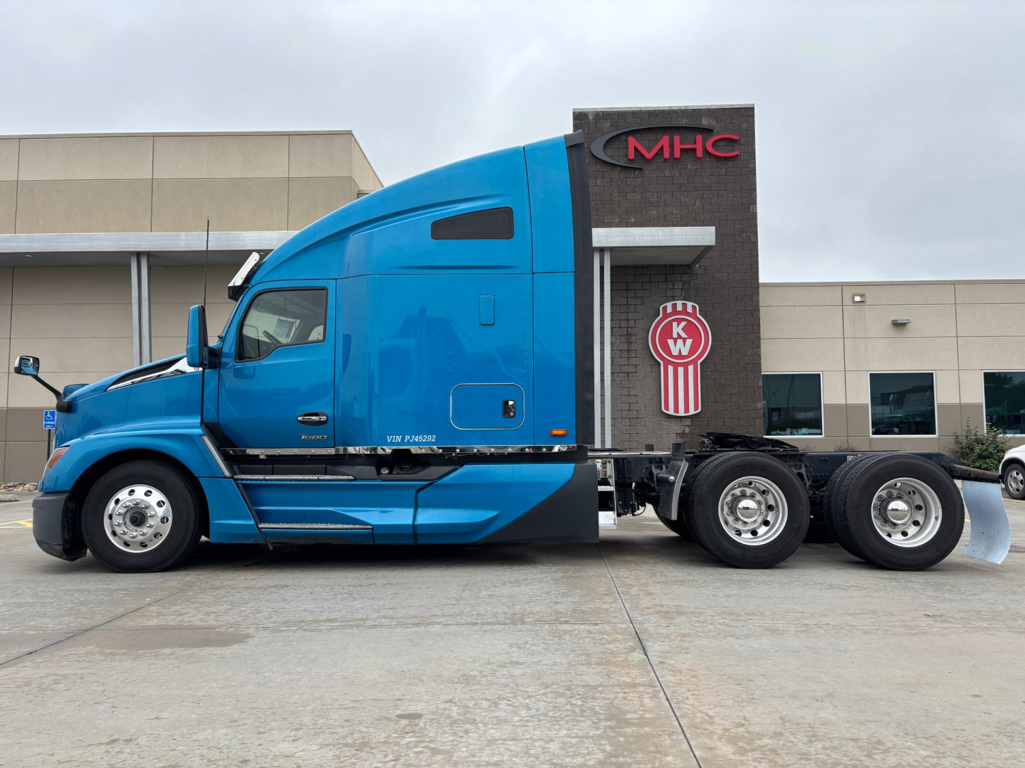 Kenworth T680 tractor unit in blue side view showing sleeper cab and three rear axles with MHC logo on building background.