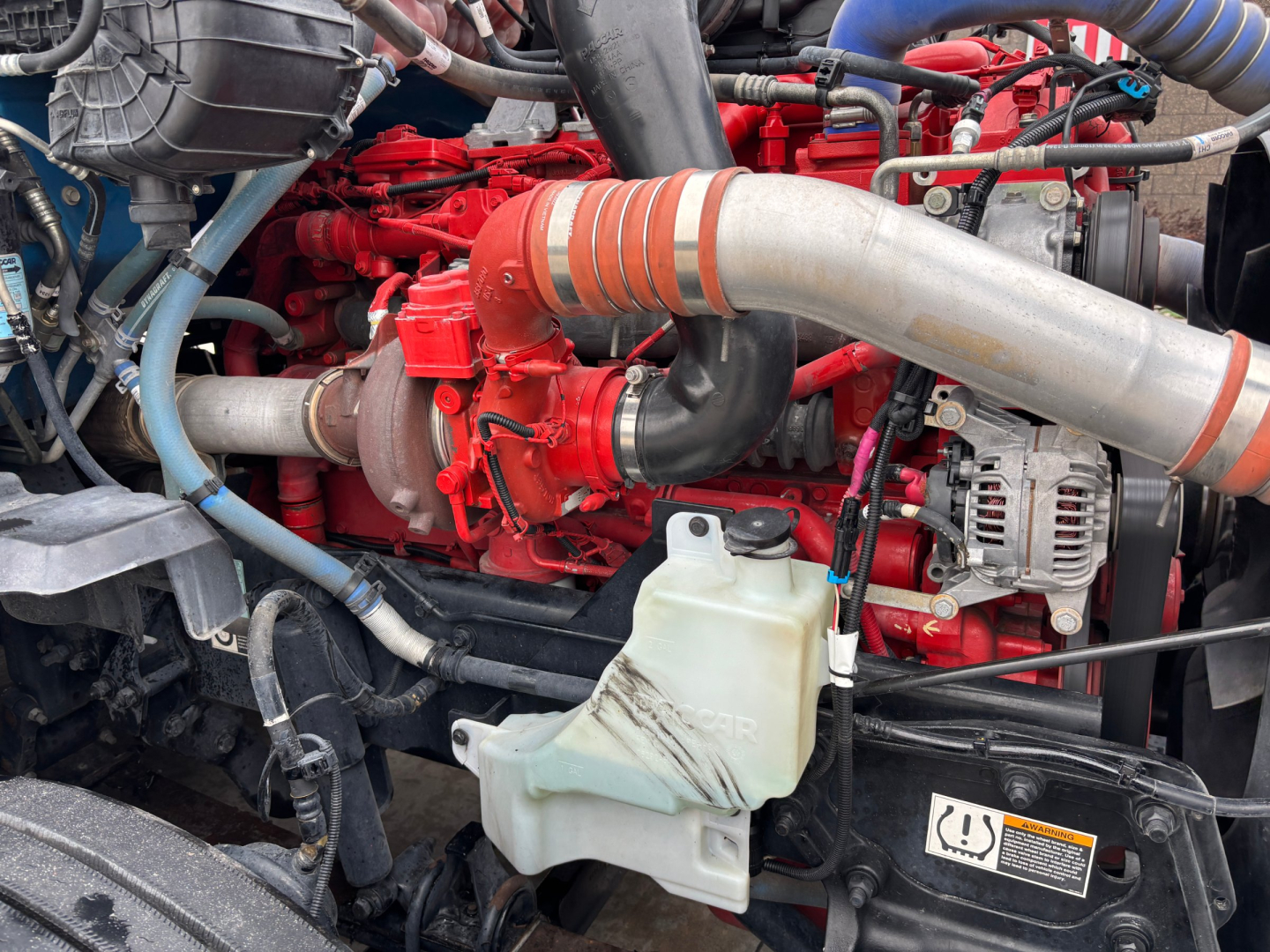 Kenworth T680 engine bay showing red engine, large silver exhaust pipe, coolant reservoir, hoses, and alternator.