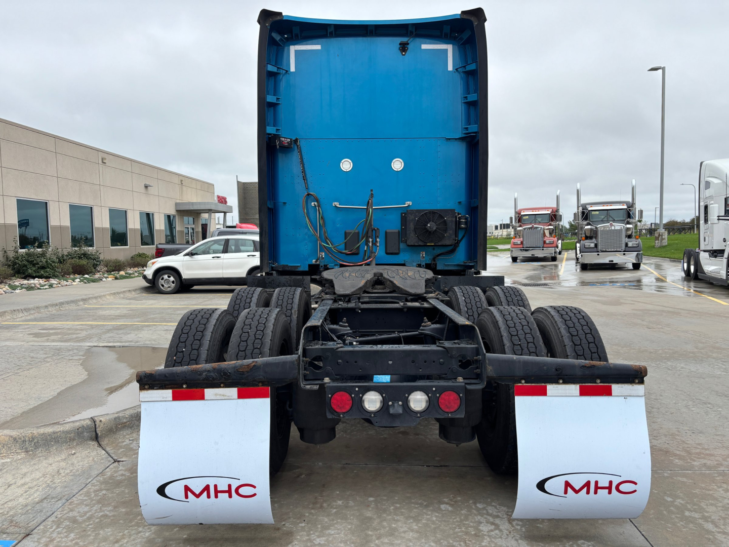 Kenworth truck rear view showing blue sleeper cab and dual rear axles with mud flaps and MHC logo on the white guards.