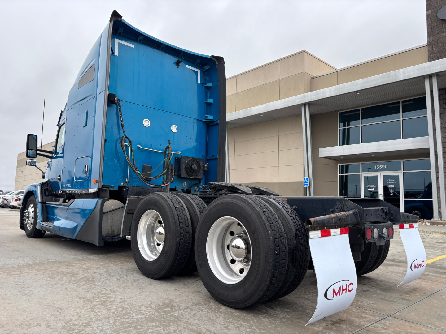 Kenworth T680 sleeper tractor chassis rear view showing wheels and blue cab, rear mudflaps with MHC logo.