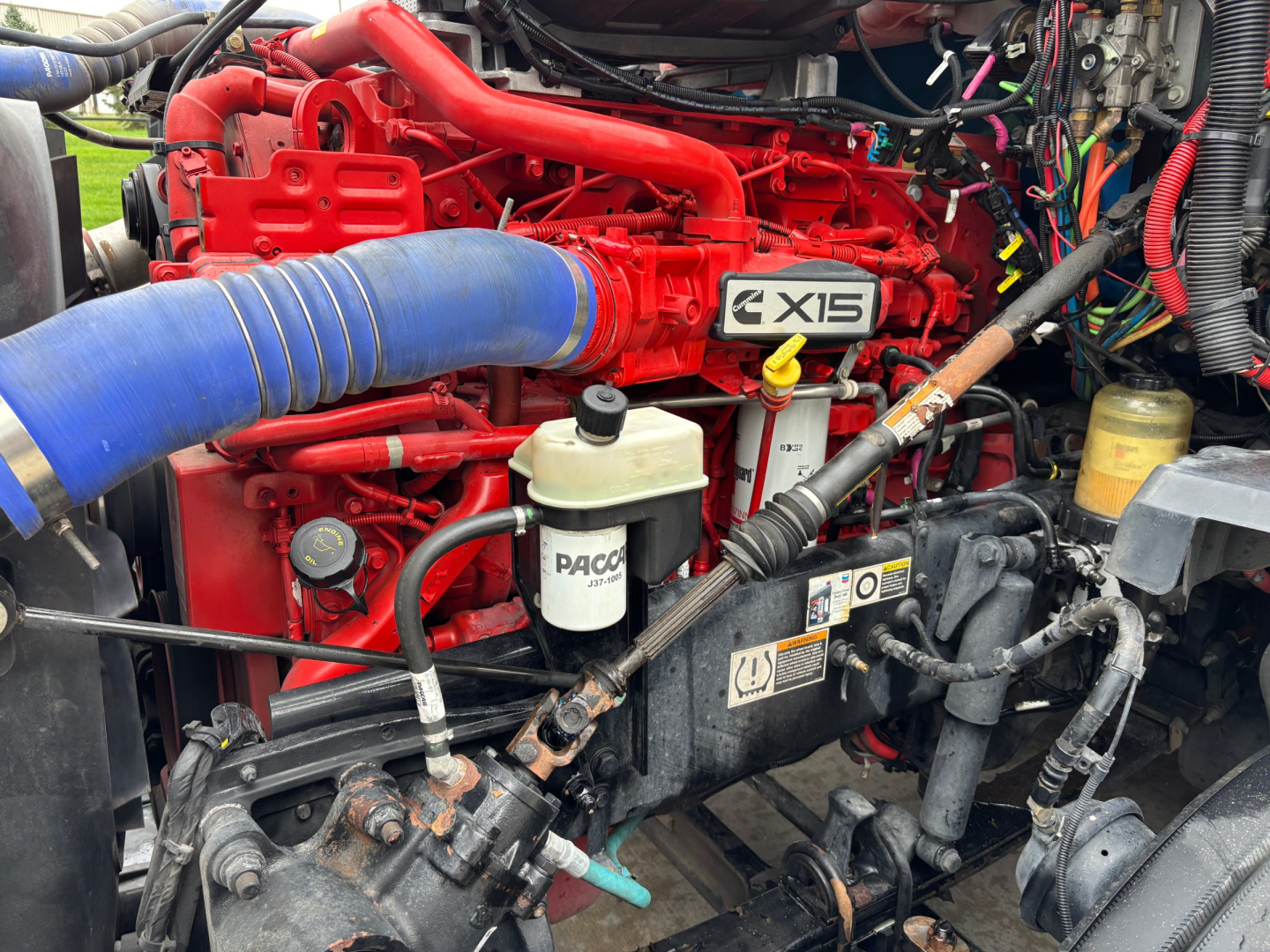 KENWORTH T680 engine bay showing red V12? no, red X15 engine with blue intake hose, white PACCAR fluid reservoir, various hoses and wiring.