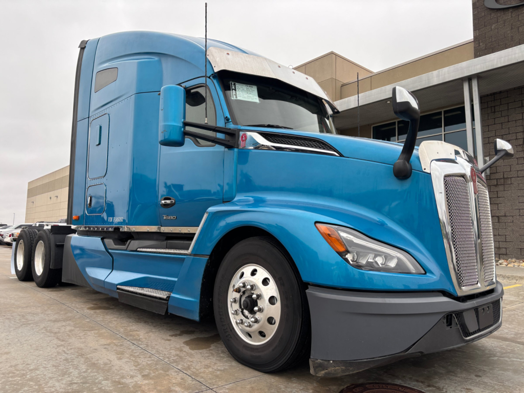 Kenworth T680 tractor in blue showing front and side. Visible text includes no legible brand or model text on image itself besides vehicle design. Scene shows a blue semi-truck with sleeper cab and chrome grille.