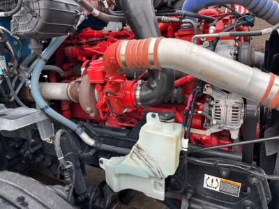 Kenworth T680 engine bay showing red engine, large silver exhaust pipe, coolant reservoir, hoses, and alternator.