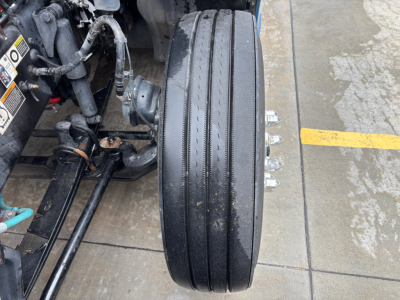 Kenworth T680 front wheel assembly and suspension components visible with tire and hydraulic lines. Flat tread tire on drive axle.