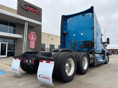 Kenworth T680 tractor rear/side view showing fifth wheel, dual rear axles, blue sleeper cab, MHC dealership building in background.