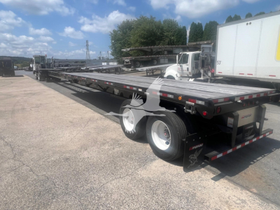 Manac flatbed trailer with extendable gooseneck at rear, dual rear axles, and reflective safety markings; side rails and long steel deck visible.