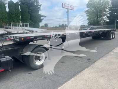 Manac flatbed trailer with extendable middle section, triple-axle rear, and black chassis. Side rails and reflective markers visible along the frame.