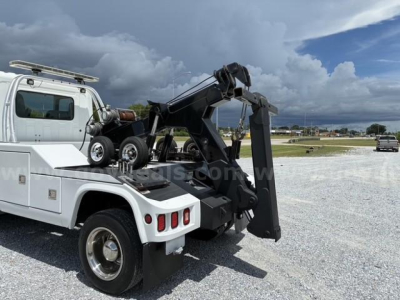 Tow truck with a front-loading hooklift system and crane in the bed, white cab, on a gravel lot with a cloudy sky.