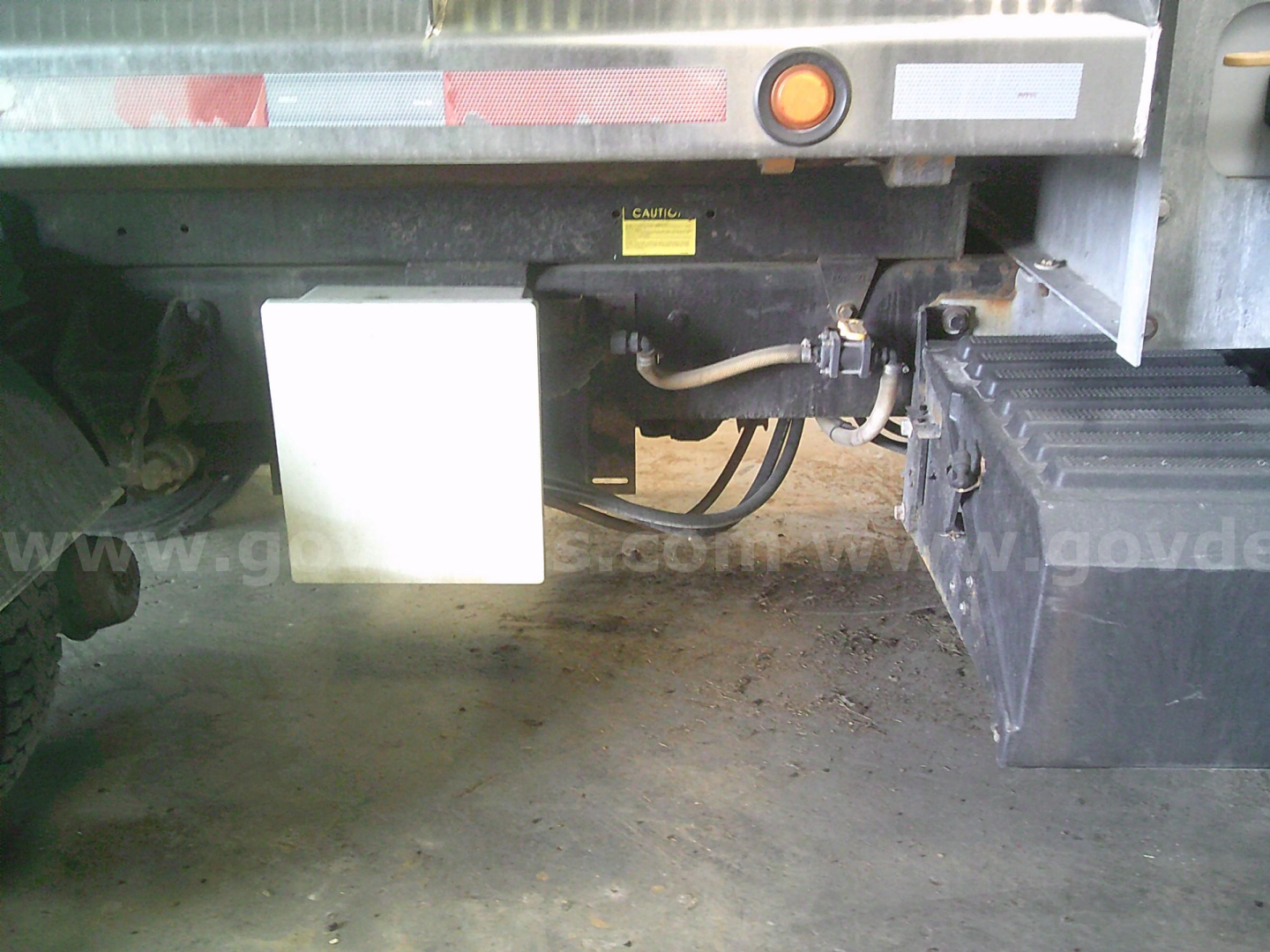 Truck rear undercarriage showing fuel/DEF lines and white rectangular container with black toolbox on right side