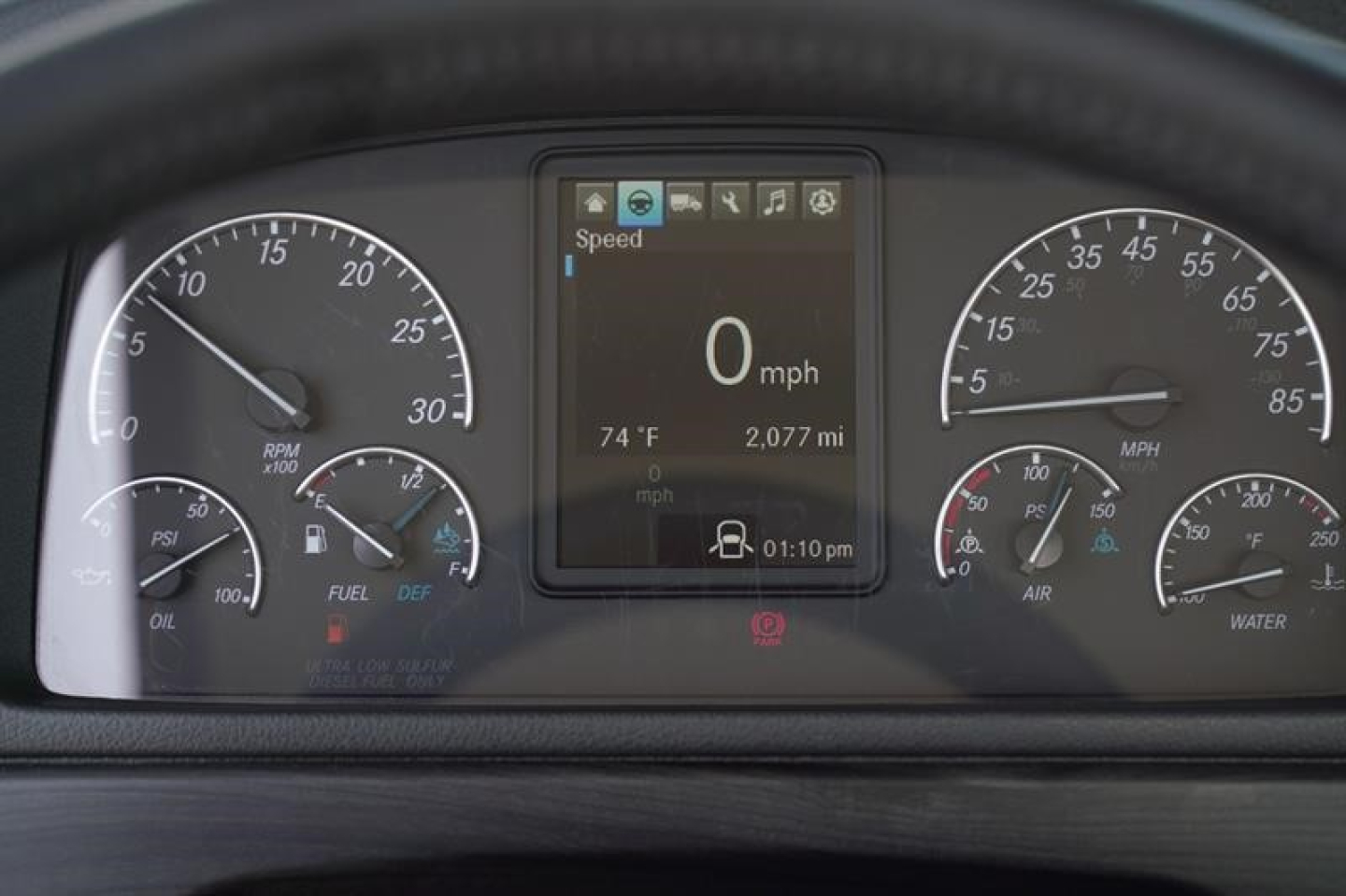 Dashboard cluster of a Freightliner M2 106 Plus showing speedo, tachometer, center digital display reading 0 mph, 74 F, 2,077 mi, time 01:10 pm.
