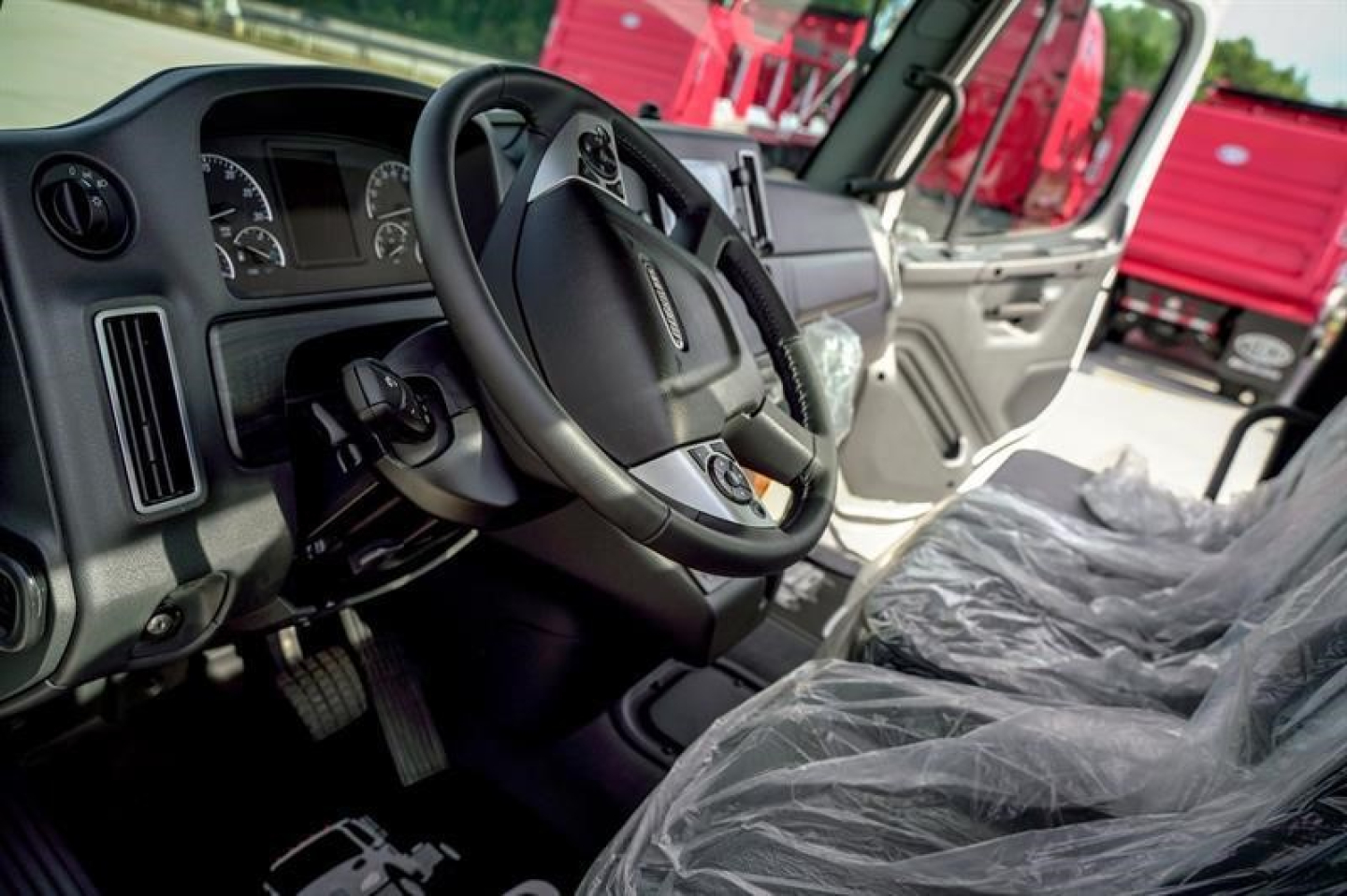 Cab interior of a Freightliner M2 106 Plus showing steering wheel, instrument cluster, and plastic-covered seats; dashboard and open door visible; exterior red trucks in background.