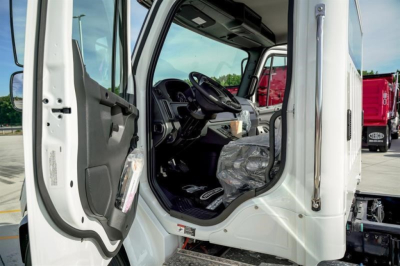 Freightliner M2 106 Plus cab interior showing steering wheel and driver seat; open door with dashboard and entry step visible.