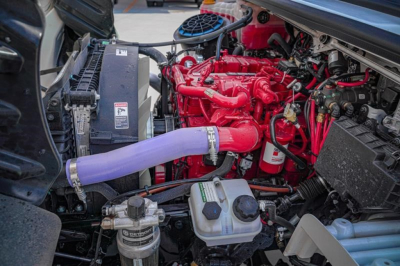 Engine bay of Freightliner M2 106 Plus showing red engine, purple coolant hose, radiator, and fluid reservoirs.