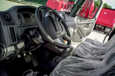 Cab interior of a Freightliner M2 106 Plus showing steering wheel, instrument cluster, and plastic-covered seats; dashboard and open door visible; exterior red trucks in background.