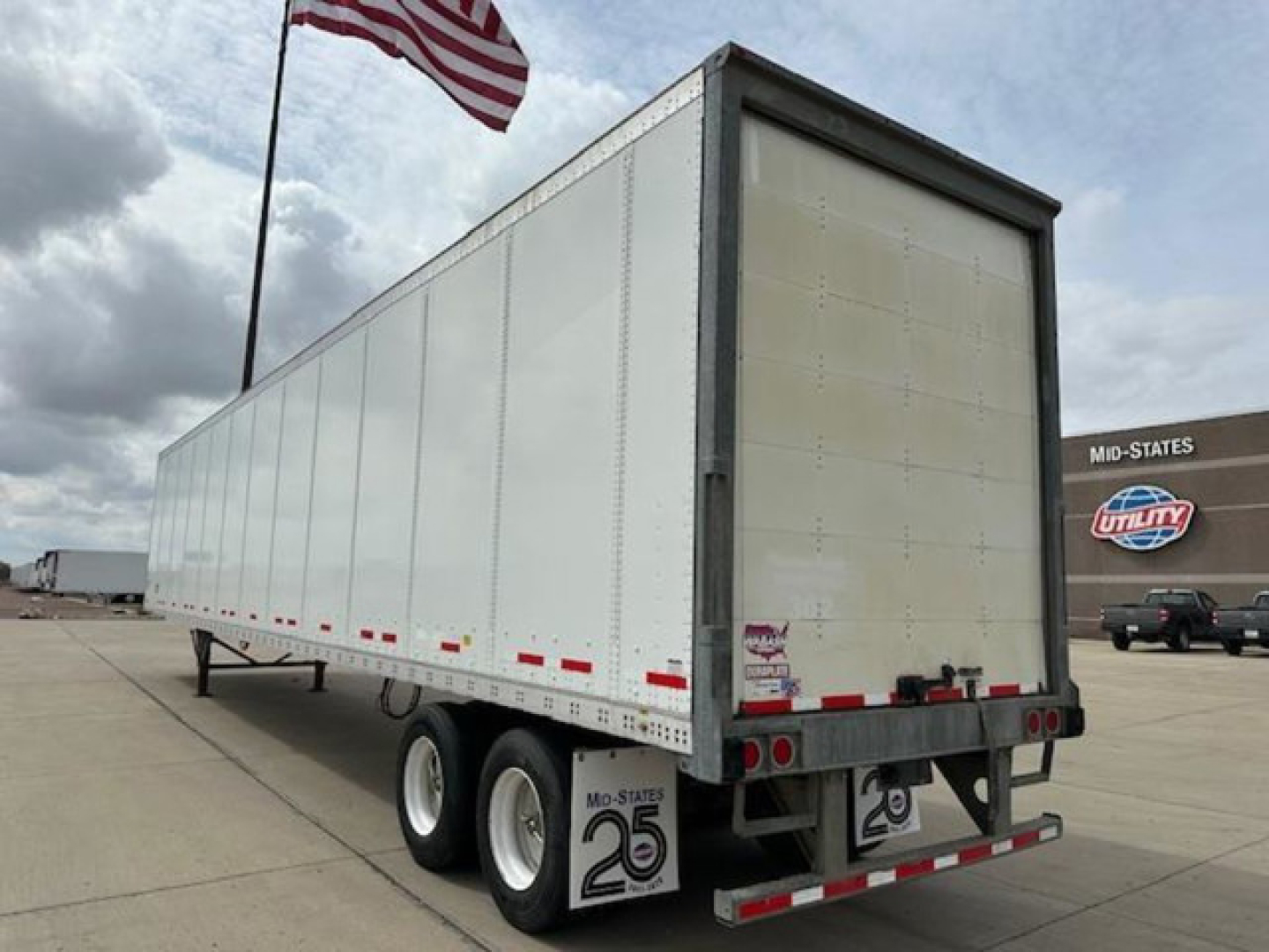 Trailer rear and side view showing enclosed white box trailer with dual rear doors, mud flaps, and US flag in background.
