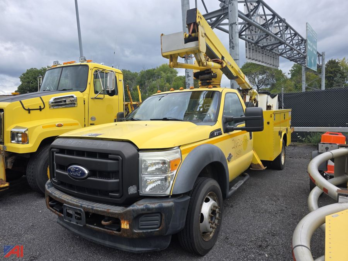 Ford F-550 bucket truck with yellow utility body and mounted aerial lift, front view showing Ford grille and bucket boom near cab; orange safety cones and other equipment in lot.