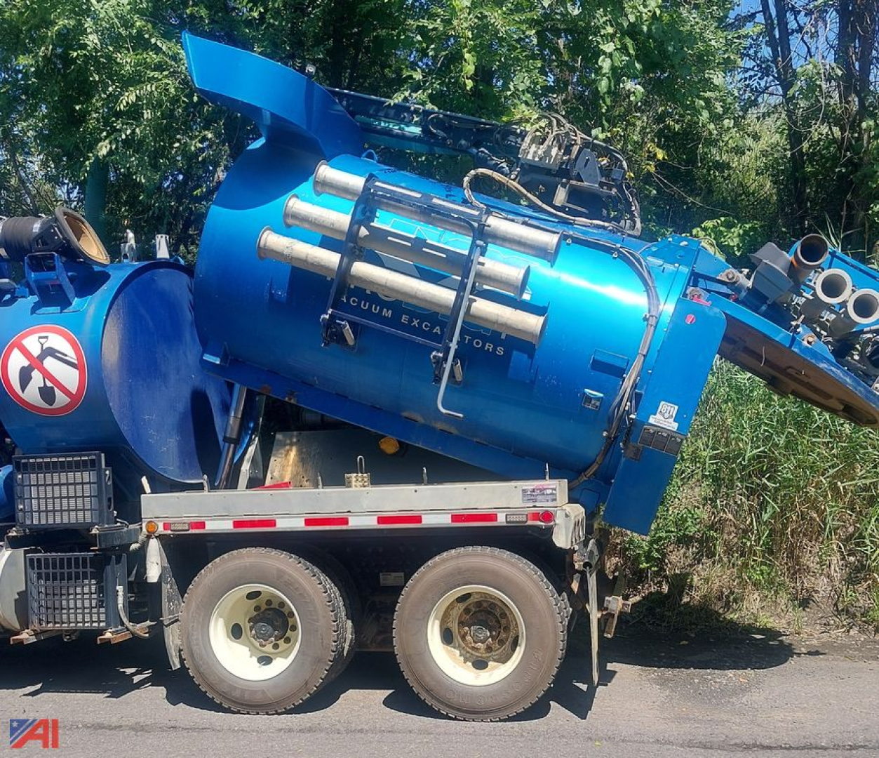 Blue vacuum tank on a dual-axle trailer with piping and control hoses; side shows text 'VACUUM EXCAVATORS' on a cylindrical body and some labels; background trees.