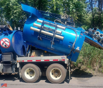 Blue vacuum tank on a dual-axle trailer with piping and control hoses; side shows text 'VACUUM EXCAVATORS' on a cylindrical body and some labels; background trees.