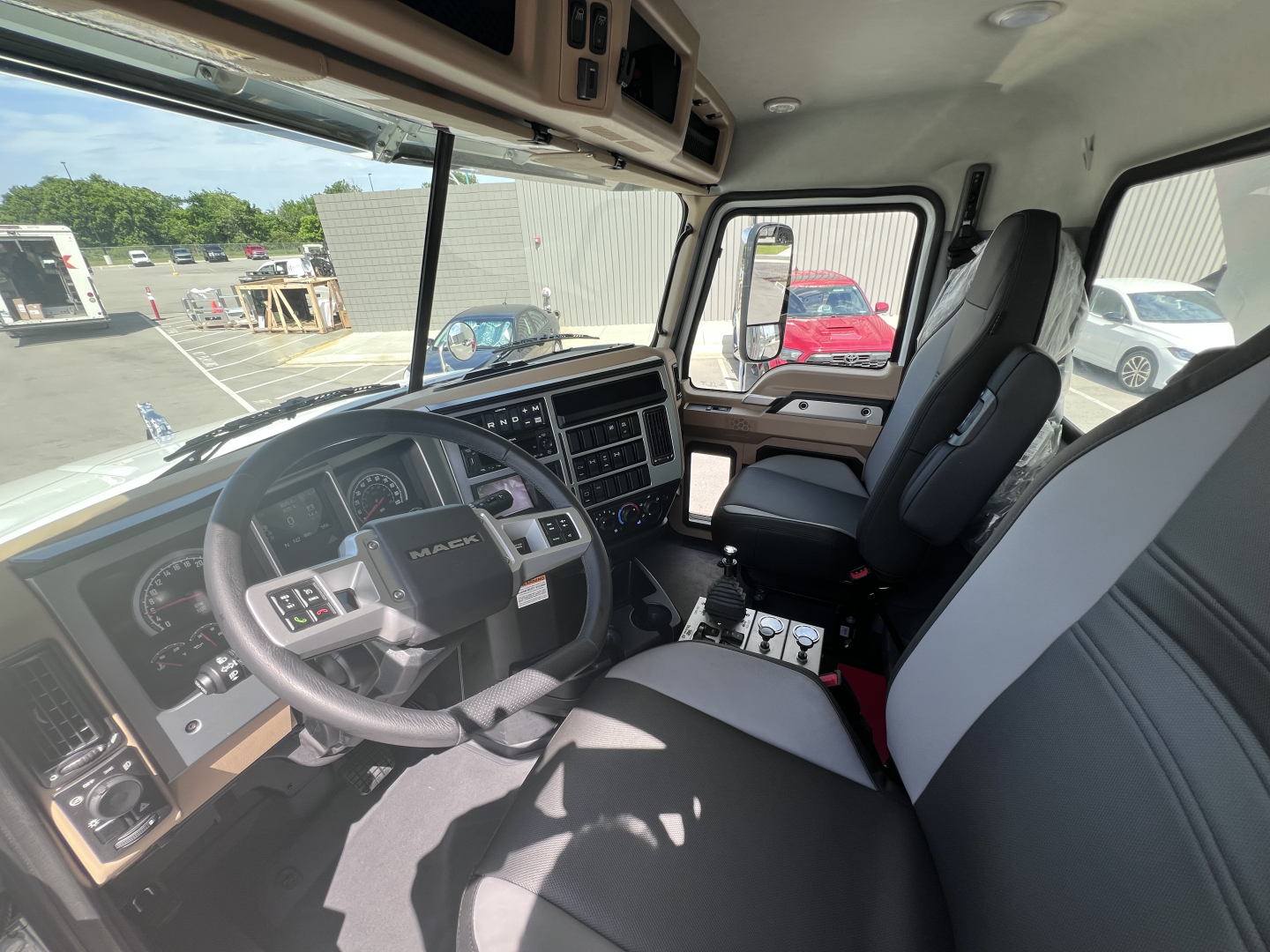 Mack GR64F cab interior showing steering wheel, dashboard with multiple gauges, and driver seat; front passenger seat and center console visible; light gray/black interior with wood trim.