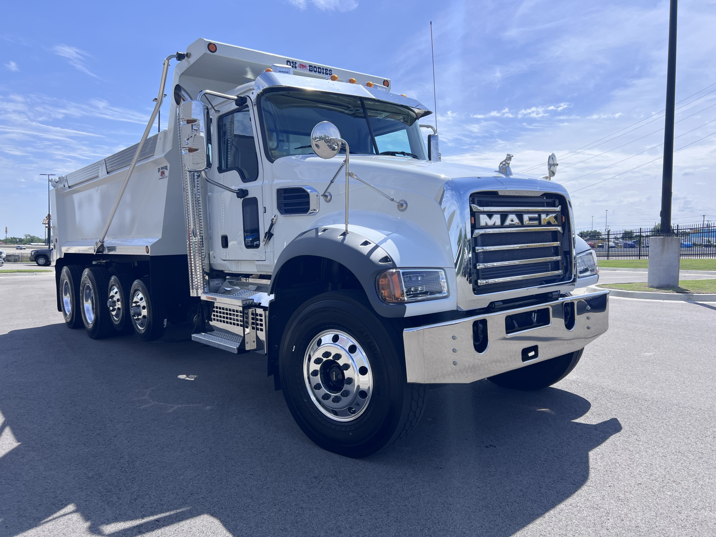 Dump truck in blue sky showing front grille with MACK badge and heavy chrome bumper, dual front wheels, and white cab with dump bed.