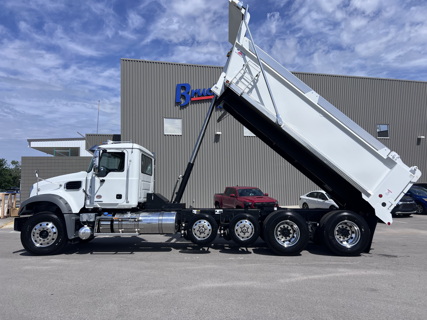 Truck chassis with dump body raised, side view of a Mack GR64F dump truck in front of a dealership.