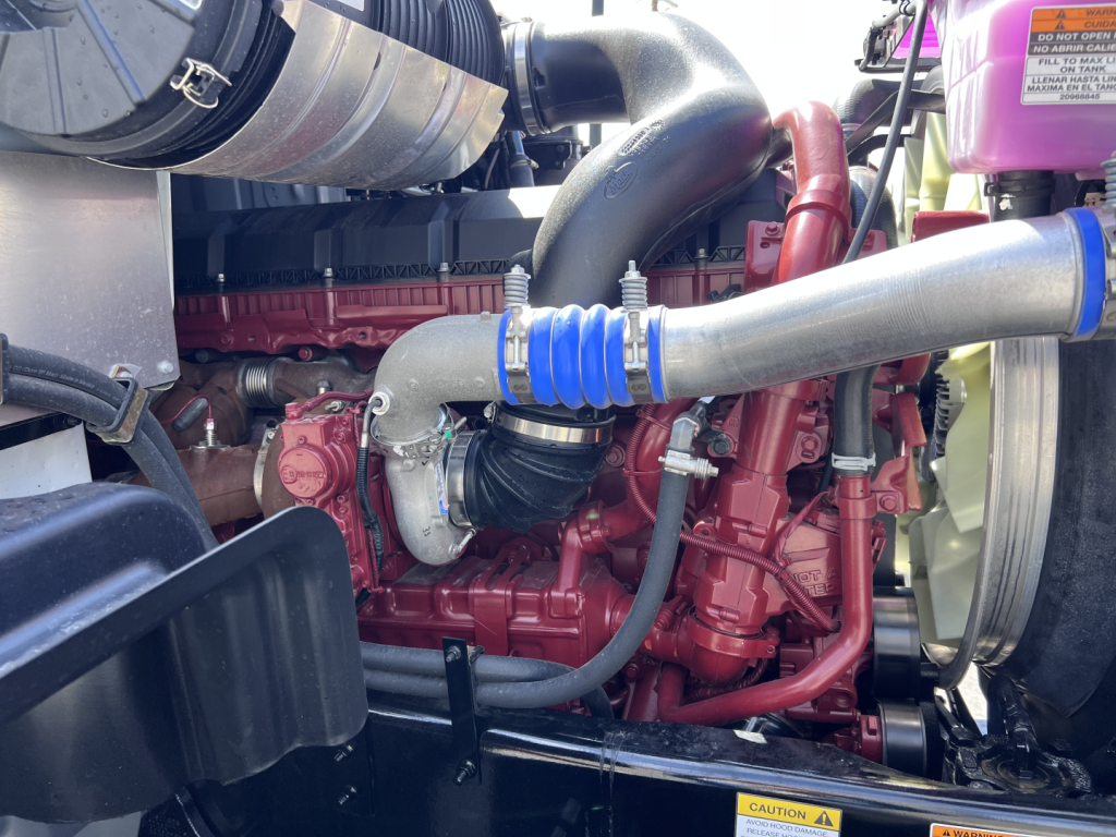 Mack engine bay with red diesel engine, blue silicone hose, turbocharger, air intake piping, and pink coolant reservoir visible.