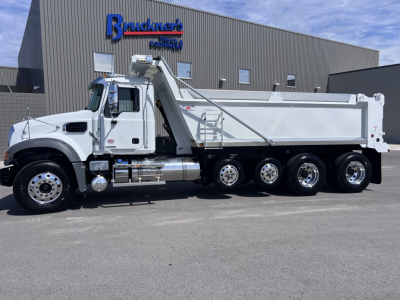 Dump truck with Rab Hi-rail Mack GR64F chassis in front of a dealership, white cab, large dump bed, multiple axles, chrome exhaust and fuel tanks, branding on building.