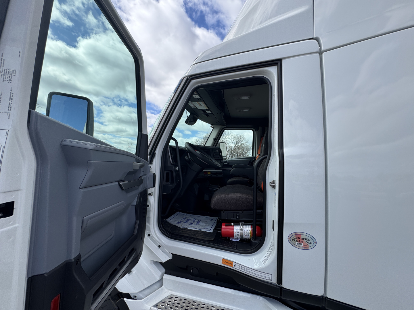Truck cab interior visible through open door of white 2026 Mack PR64T. Dashboard, steering wheel, and driver seat seen; door panel with manual window crank; exterior mirrors and side step indicate a new conventional daycab.