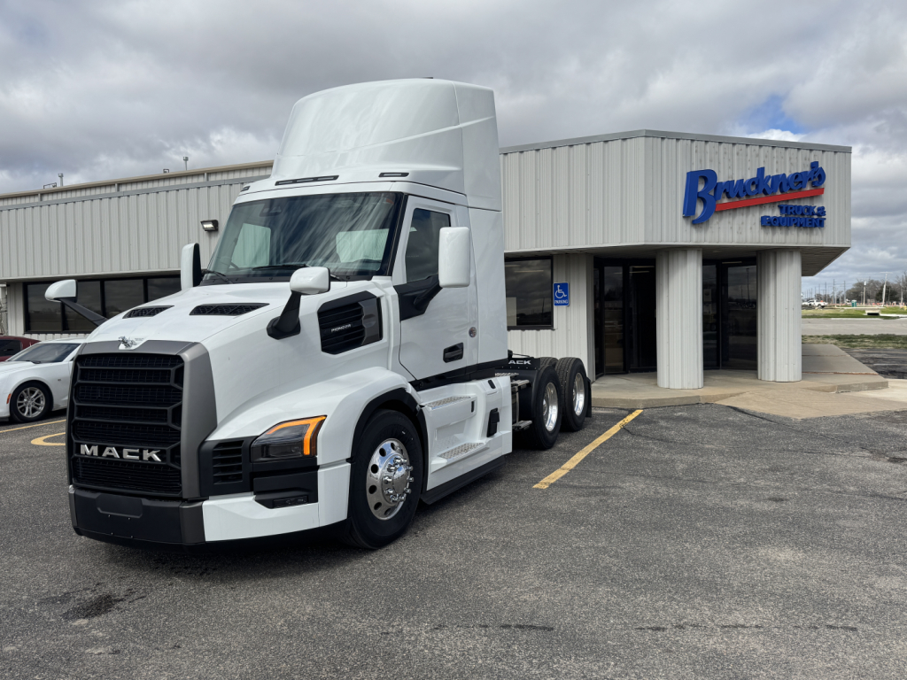 Mack PR64T tractor (cab and front) in white, 3-axle configuration, in front of a dealership building with Mack logo visible.