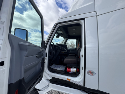 Truck cab interior visible through open door of white 2026 Mack PR64T. Dashboard, steering wheel, and driver seat seen; door panel with manual window crank; exterior mirrors and side step indicate a new conventional daycab.