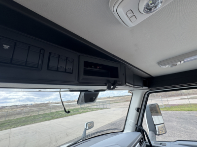 Cockpit view of a new 2026 Mack PR64T truck showing overhead storage compartments and interior controls. Visible text includes labels on the overhead panel and rearview mirror area.