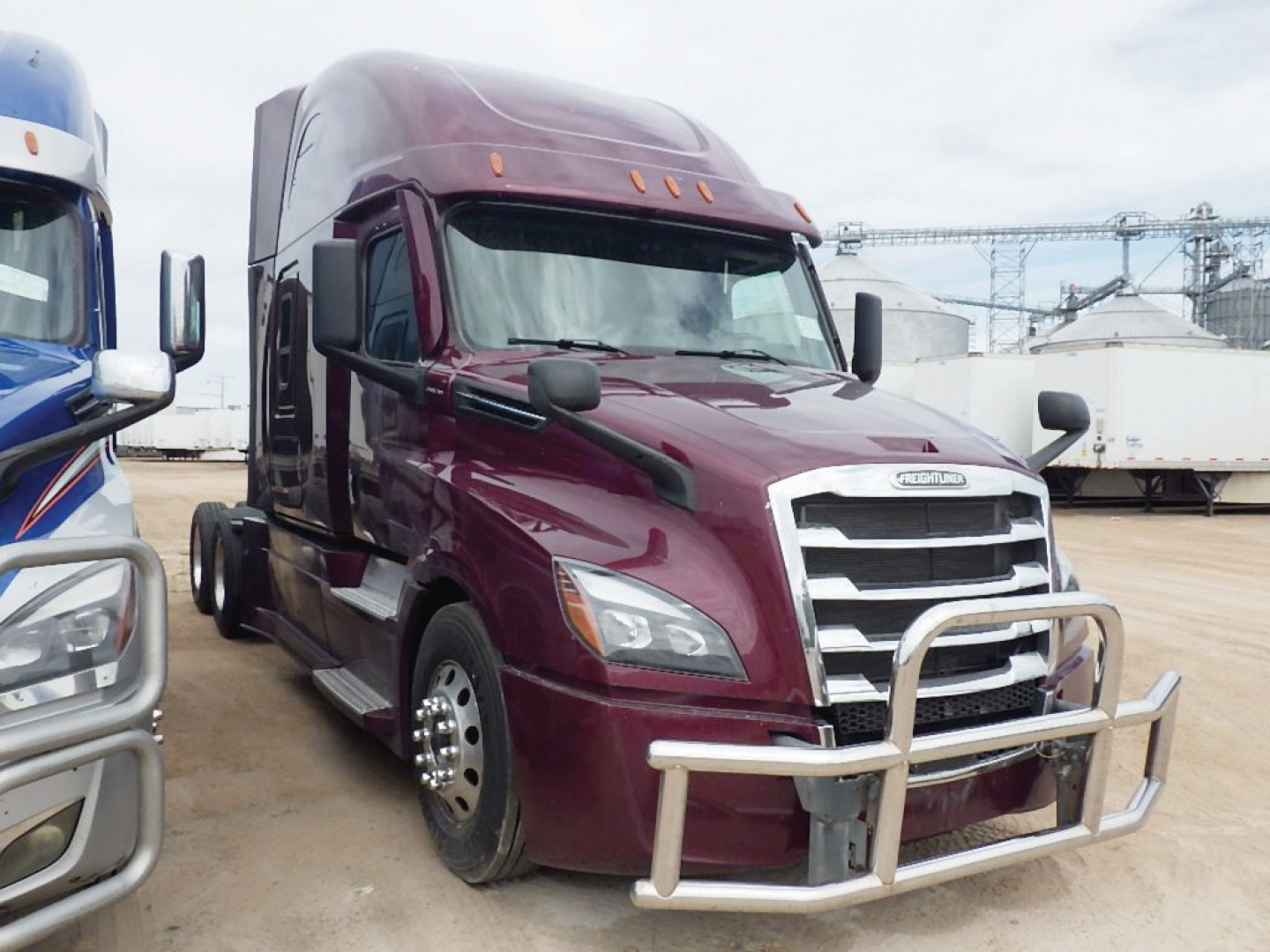 Freightliner Cascadia126 sleeper cab shows maroon exterior with large chrome grille and front bull bar. Left side shows dual rear wheels; other trucks and industrial background.